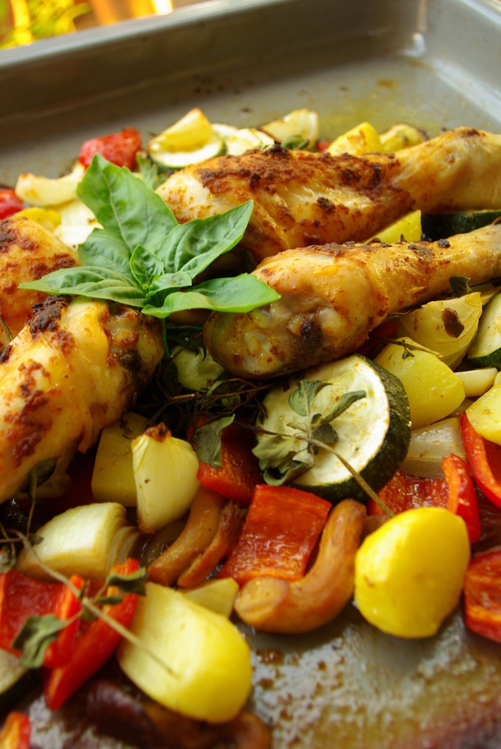 Hähnchenkeule auf Gemüsebett. Buntes Gemüse wie rote Paprika, gelbe Kartoffeln, weiße Zucchini, orangen Karotten auf einem Backblech. Vier Hähnchenkeulen obendrauf, Garnitur zwei Basilikum.