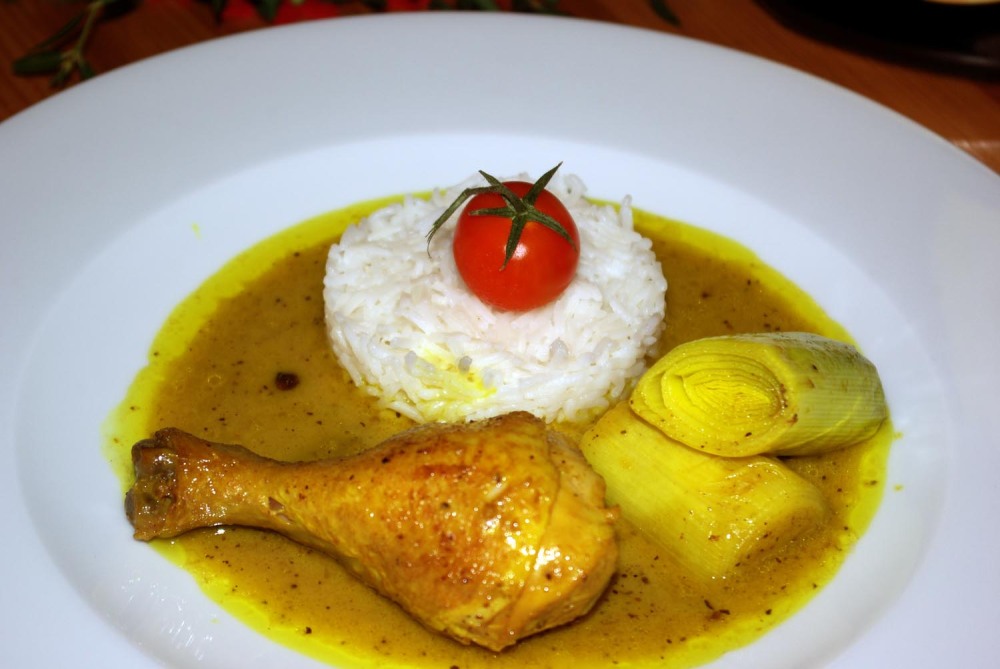 Vergoldetes Hähnchen mit Basmatireis und Lauch. Weißer Teller, Hähnchenkeule, 2 Stücke grüner Lauch, rund angerichtet Basmatireis, darauf eine rote Cocktailtomate.