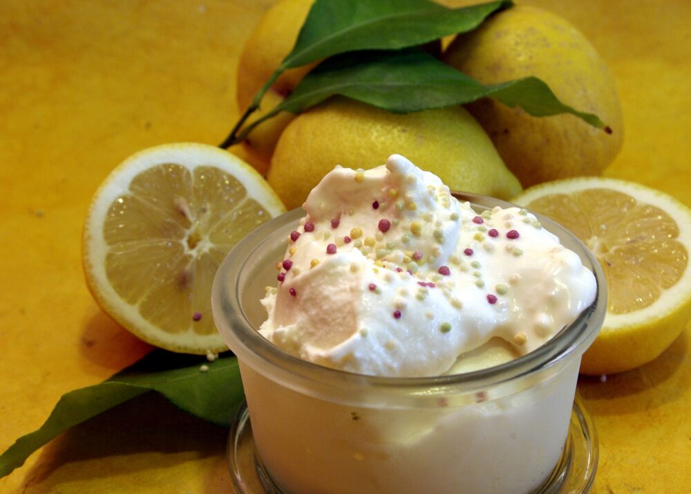 Zitroneneis mit Schwedenmilch und Zitronenmarmelade. WEckglas gefüllt mit Zitroneneis, darauf Schlagsahne. Im Hintergrund frische Zitronen mit Blättern.