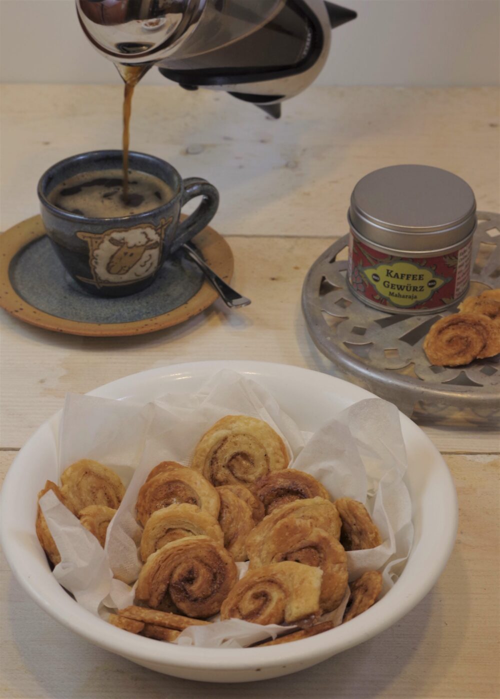 Blätterteigschnecken mit KaffeeFee. Im Vordergrund steht ein tiefer Teller mit den Blätterteigschnecken, dahinter wird gerade aus einer kleinen French-Press-Kanne Kaffee in eine Espressotasse geschüttet.