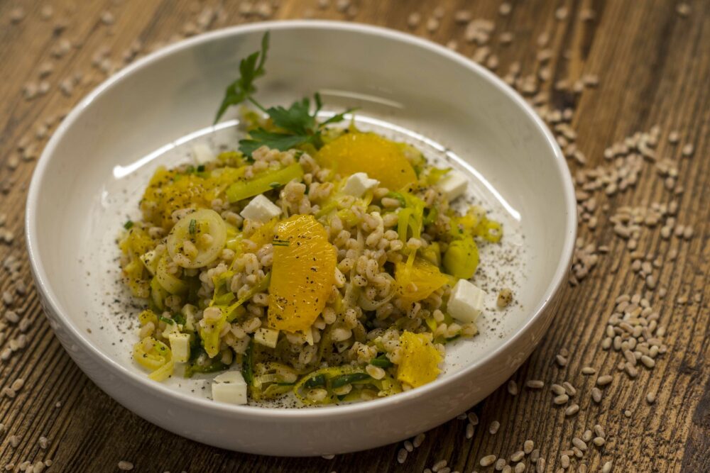 Dinkelreis-Salat mit Lauch und Vadagam Vadouvan. Auf einem tiefen beigen Teller wurde der Dinkelreis,vermischt mit Lauchstreifen angerichtet. Eine Garnitur aus Petersile und Orangenfilets rundet das Bild ab.