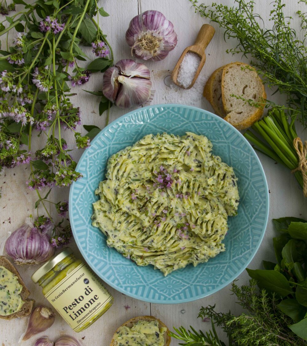 Gartenkräuterbutter - Geheimrezept aus der Geniessermanufactur. Gartenkräuterbutter mit frischen Kräutern, angerichtet auf einem türkisfarbenem Teller, umrahmt von frischen Kräutern und einem Glas Pesto di Limone.