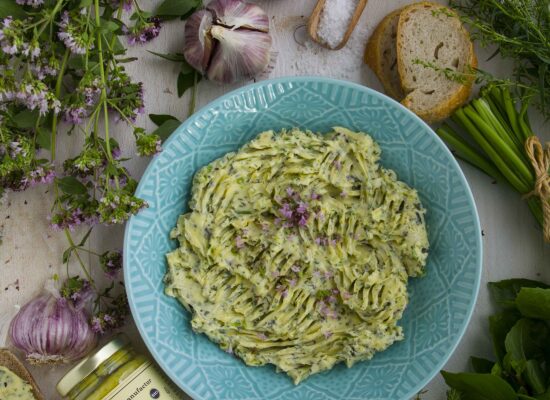 Gartenkräuterbutter - Geheimrezept aus der Geniessermanufactur. Gartenkräuterbutter mit frischen Kräutern, angerichtet auf einem türkisfarbenem Teller, umrahmt von frischen Kräutern und einem Glas Pesto di Limone.