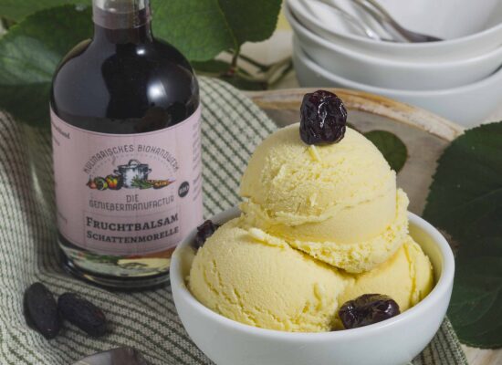 Tonkabohnen-Eis mit Schattenmorellen-Fruchtbalsam, dekoriert in einer weißen Schale, garniert mit kandierten Schattenmorellen. Im Hinergrund steht eine Flasche des Schattenmorellen-Fruchtbalsams. Der Untergrund ist eine karrierte weiß-grüne tischdecke.