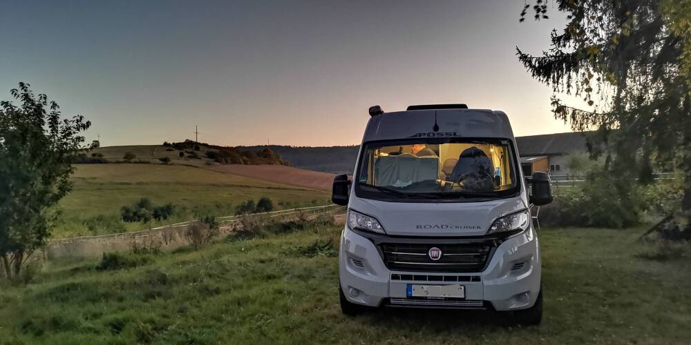 Unser erstes Mal, der Titel dieses Beitrags. Ein Campervan in Frontansicht steht auf einer Wiese, dahinter geht gerade die Sonne unter. Ein Stimmungsbild.