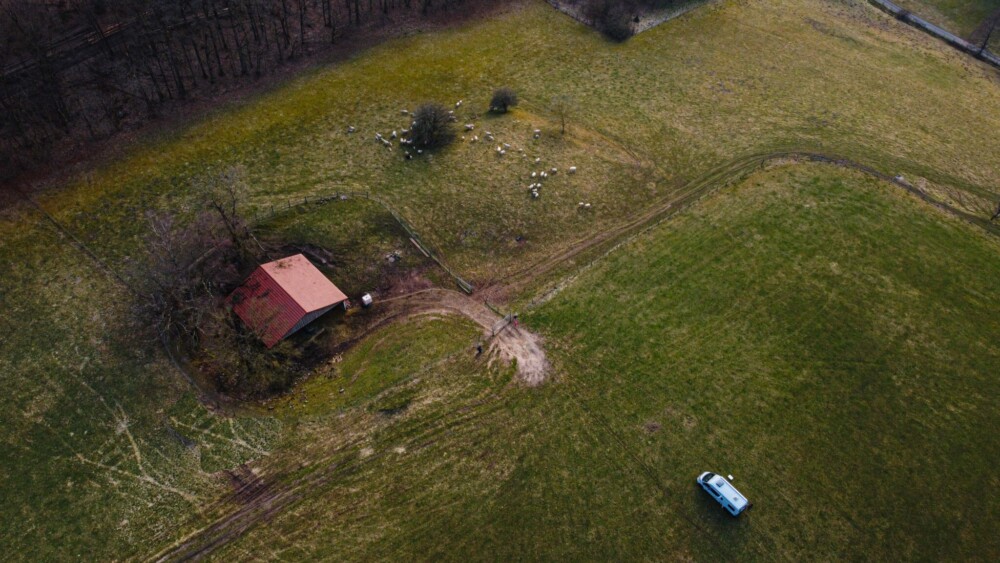 Freistehen auf dem Klebheimer Berg. Ein Luftbild. Eine große Wiese, darauf eine kleine Scheune, ein Pferch mit Schafen und ein Campervan.