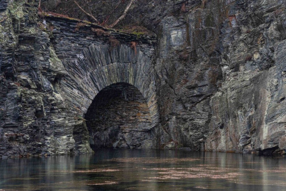 Schiefermuseum Lehesten im Thüringer Wald. Der Eingang zur Miene als Rundbogen. Davor hat sich ein See gebildet. Die Felswand ist grau und zerklüftet.