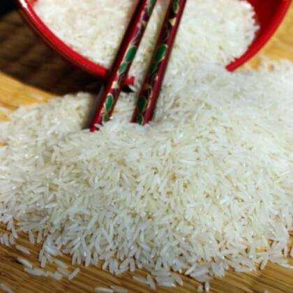 Echter Basmati-Reis Duftreis bio. Auf einer Matte liegt im Hintergrund eine rote Schale, aus der der Reis nach vorne herauszurinnen scheint. Ein paar Essstäbchen in dunklem Rot wurden quer über die Schale gelegt.