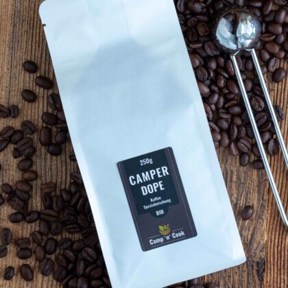 Camper-Dope Spezialmischung BIO Kaffee. Eine weiße Tüte, darum herum Kaffeebohnen und daneben ein Kaffeemaß aus Edelstahl.