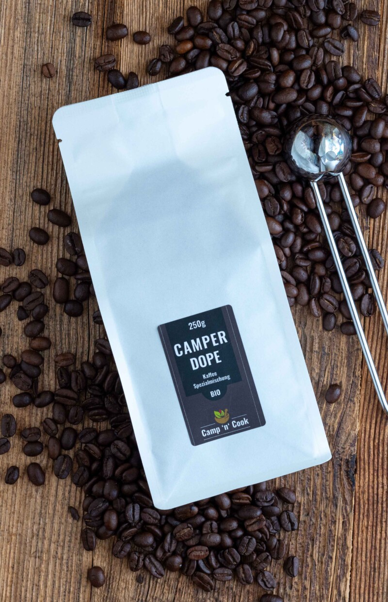 Camper-Dope Spezialmischung BIO Kaffee. Eine weiße Tüte, darum herum Kaffeebohnen und daneben ein Kaffeemaß aus Edelstahl.