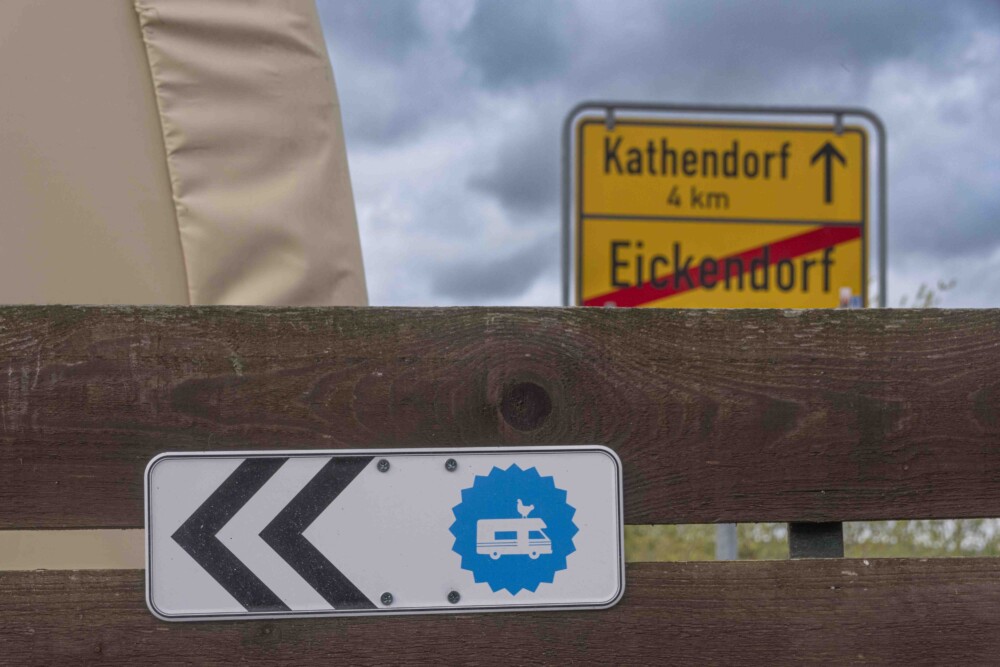 Landvergnügen Stellplatz Sue Eickendorf. Am Zaunbrett befestigt das Hinweisschild von Landvergnügen. Weißer Camper in blauem Kreis. Zwei Richtungspfeile.